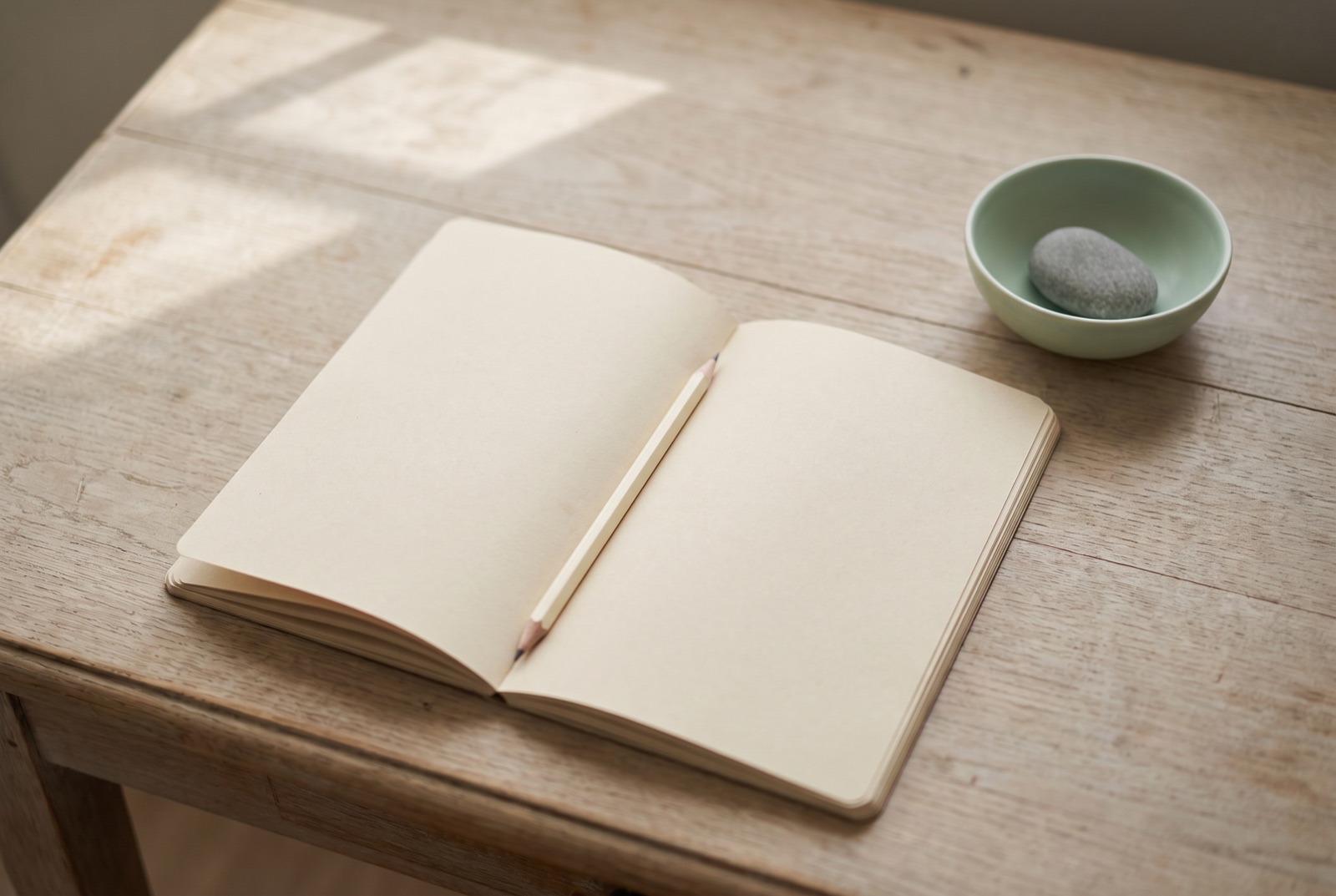 An open blank notebook with a pencil resting in the gutter on a sunlit wooden desk.