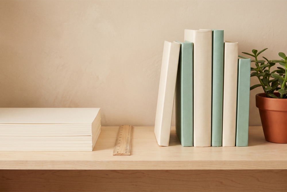 A classroom shelf with a stack of plain paper, a wooden ruler, cloth-bound books and a small plant.